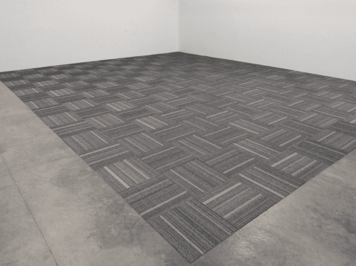 Gray carpet tiles laid in a quarter-turn pattern across a shop floor, framed by a border of bare concrete and white walls